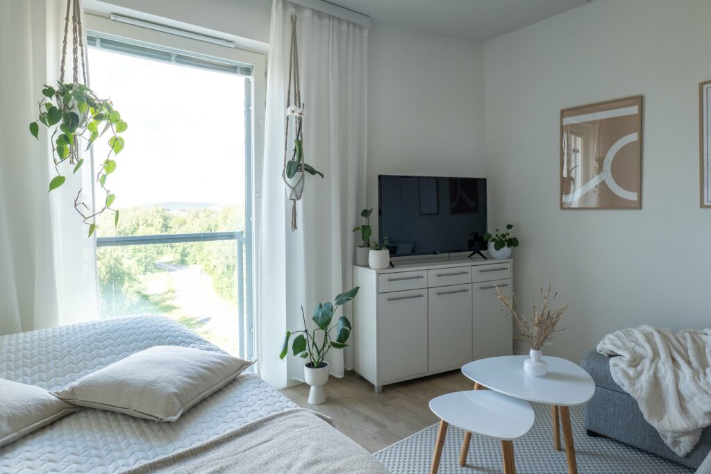Koas Kangas -kohteen asunto, näkymä huoneeseen jossa suuri ikkuna, sänky ja sen päällä pari tyynyä, senkki, jonka päällä tv sekä kaksi pientä sivupöytää keskellä lattiaa. Katosta roikkuu viherkasvi ja ulkona on aurinkoinen kesäsää.